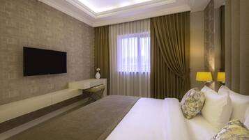 Habitación de hotel con televisión de pantalla plana en la pared, un escritorio con silla, cama con almohadas estampadas y una ventana con cortinas.