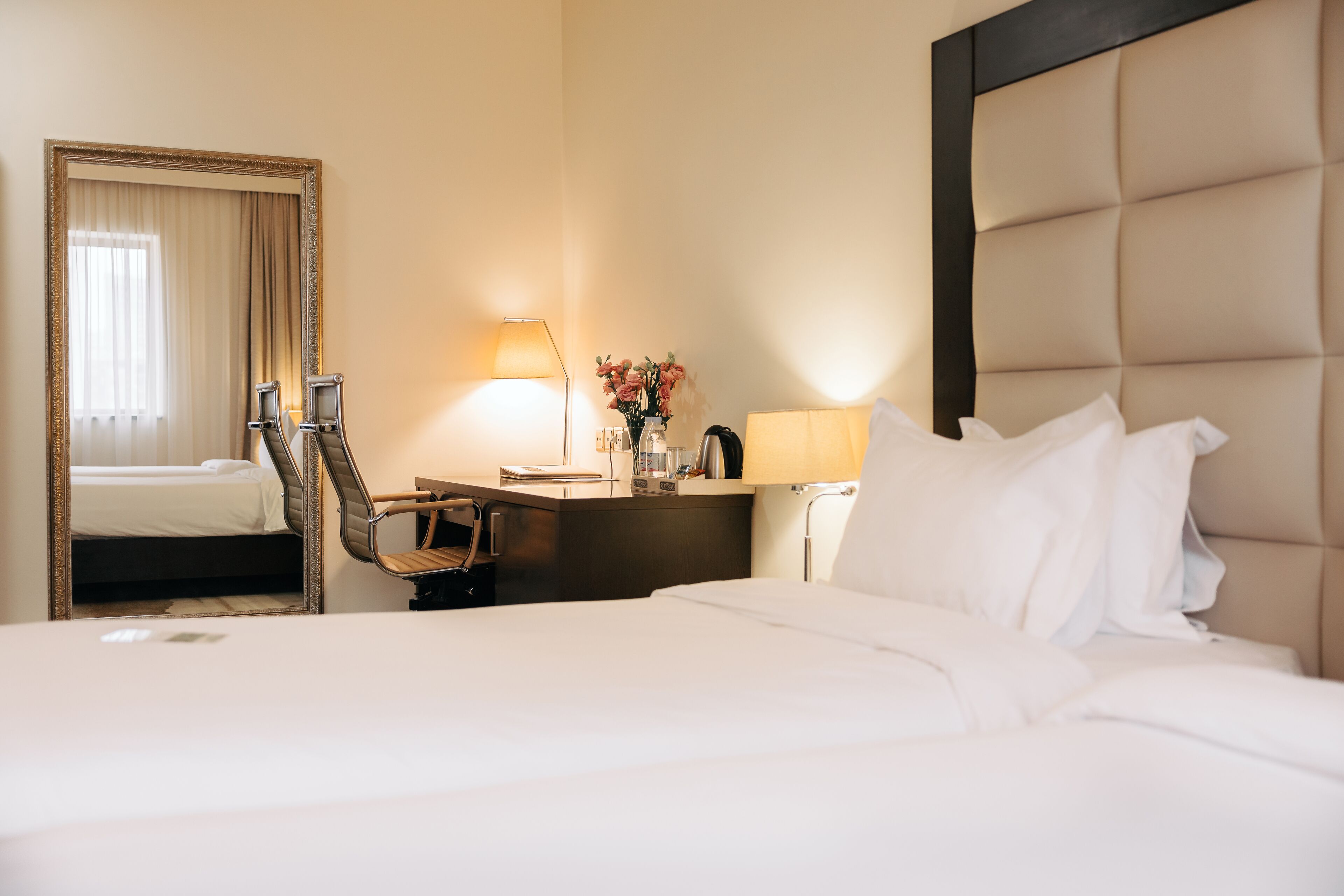 A hotel room with a large bed, a desk with a chair, a mirror, and a lamp.