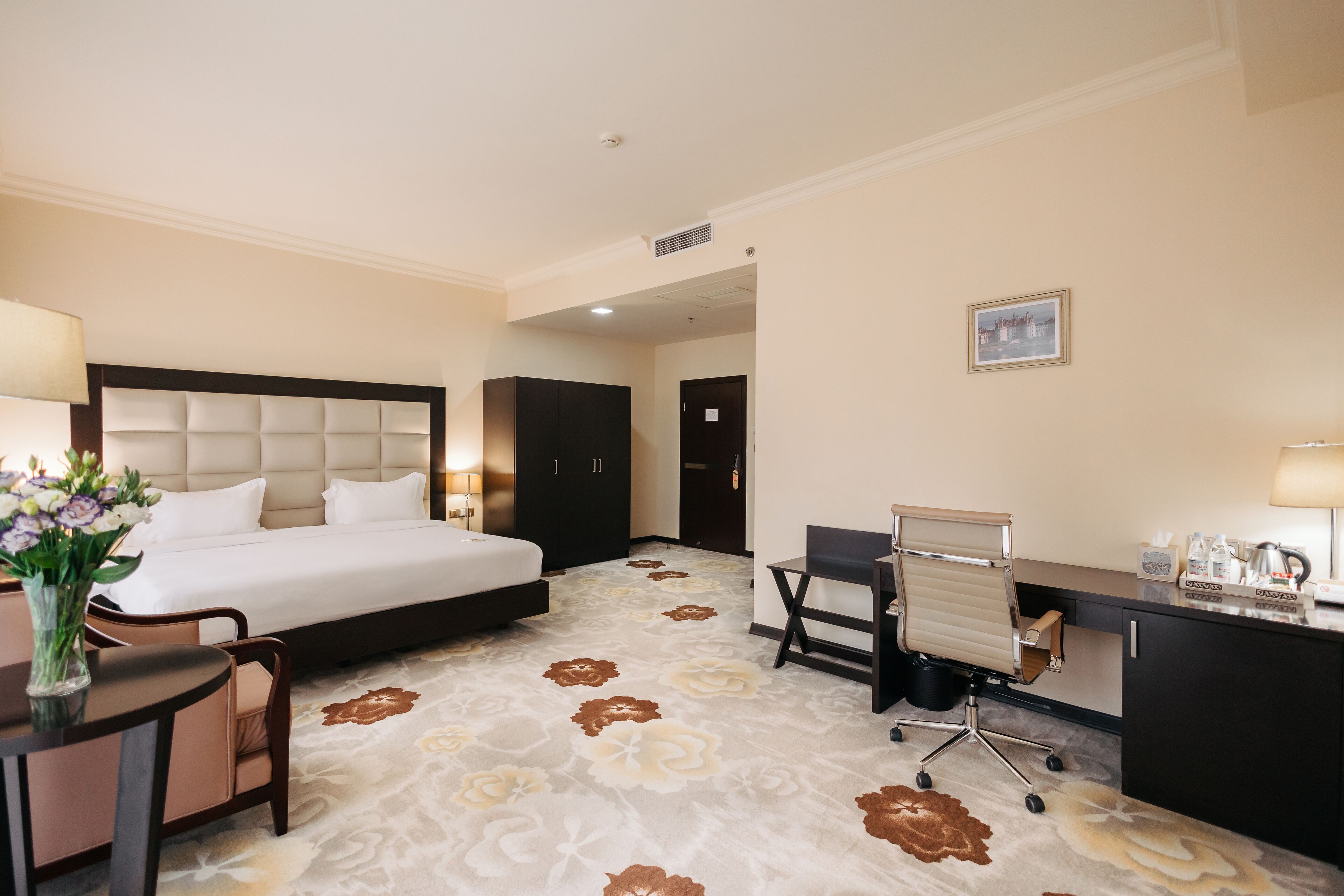 Een hotelkamer met een groot bed, een bureau met een stoel, een klein tafeltje met een bloevas en een foto aan de muur.