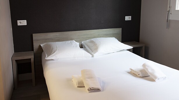 A neatly made bed with white linens and two pillows, a small wooden nightstand beside it, and a wall socket on the left. - Hotel Beaulieu (Clermont-Ferrand)