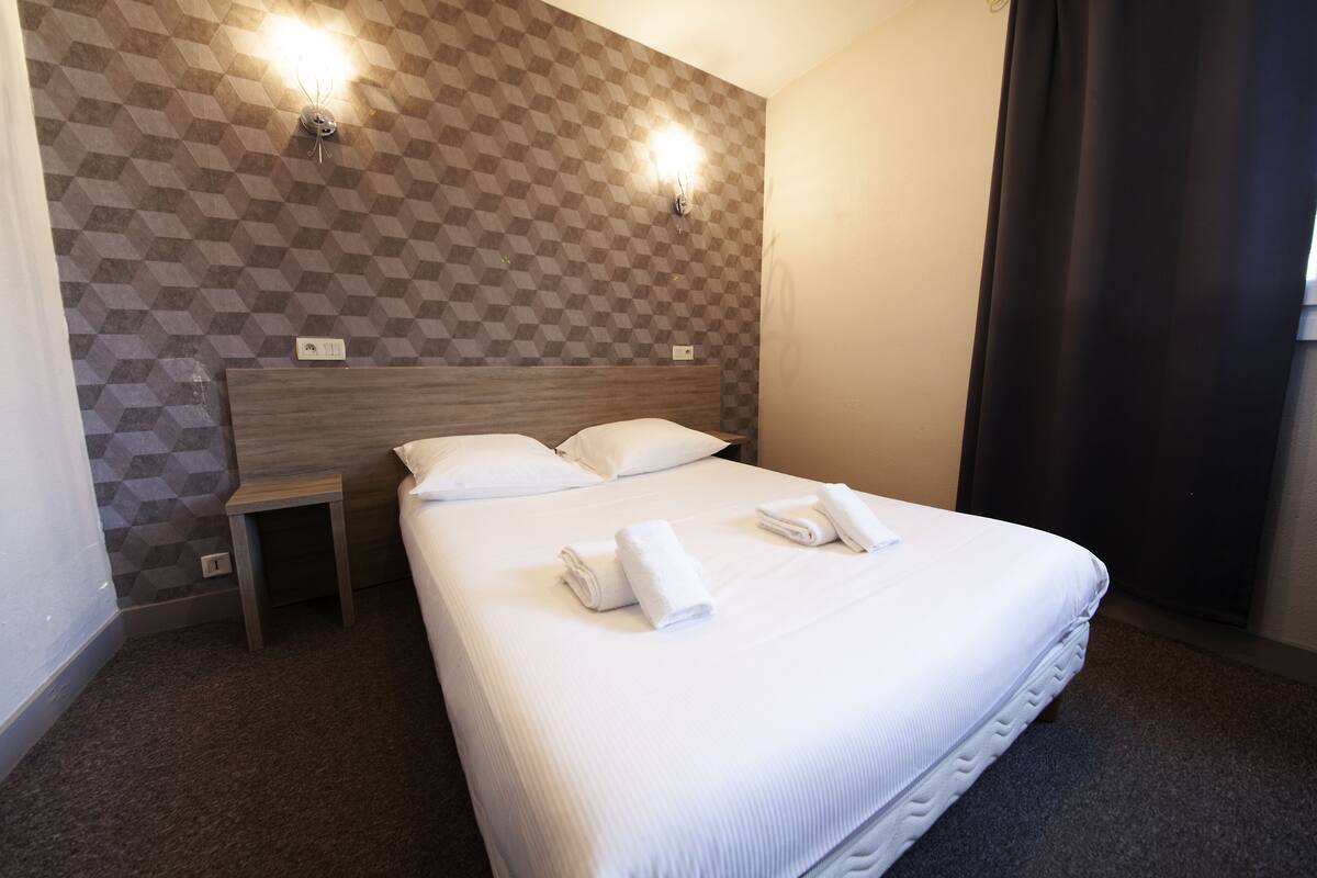 A hotel room with a checkered wall pattern, a wooden headboard, a small bedside table, and neatly folded towels on the bed.