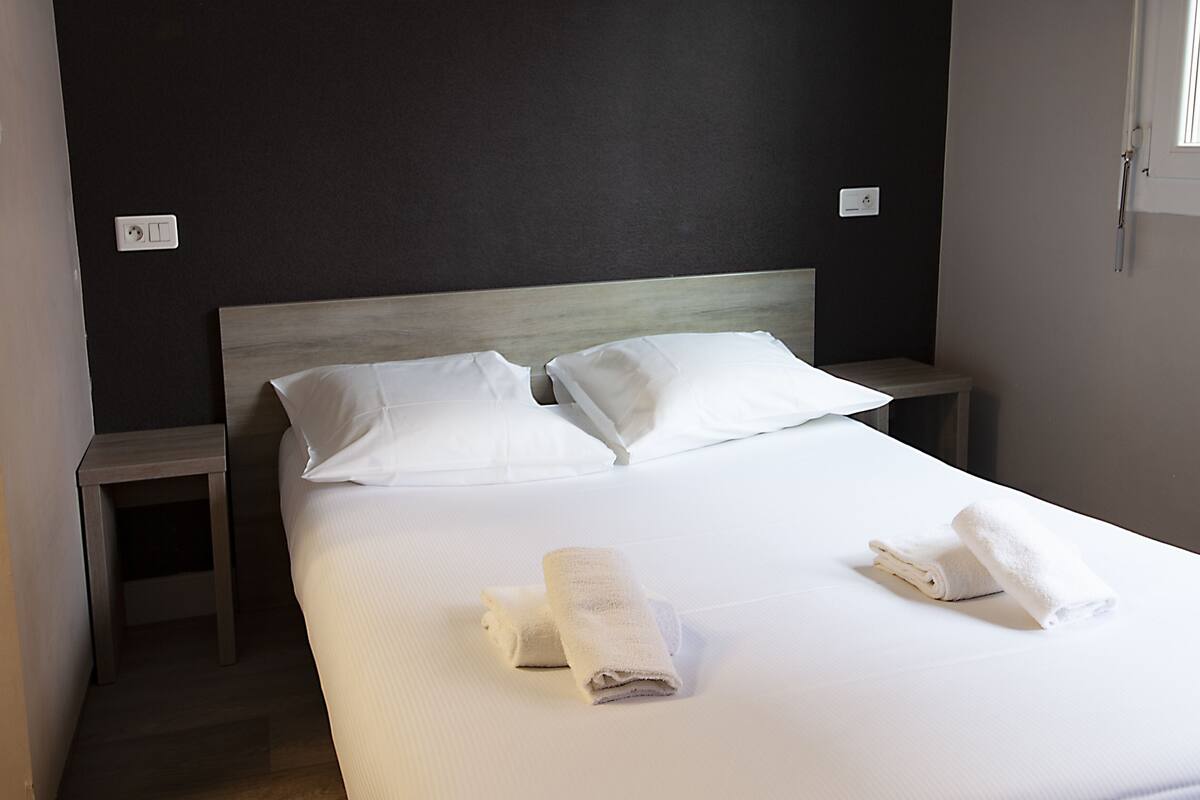 A neatly made bed with white linens and two pillows, a small wooden nightstand, and a wall socket.