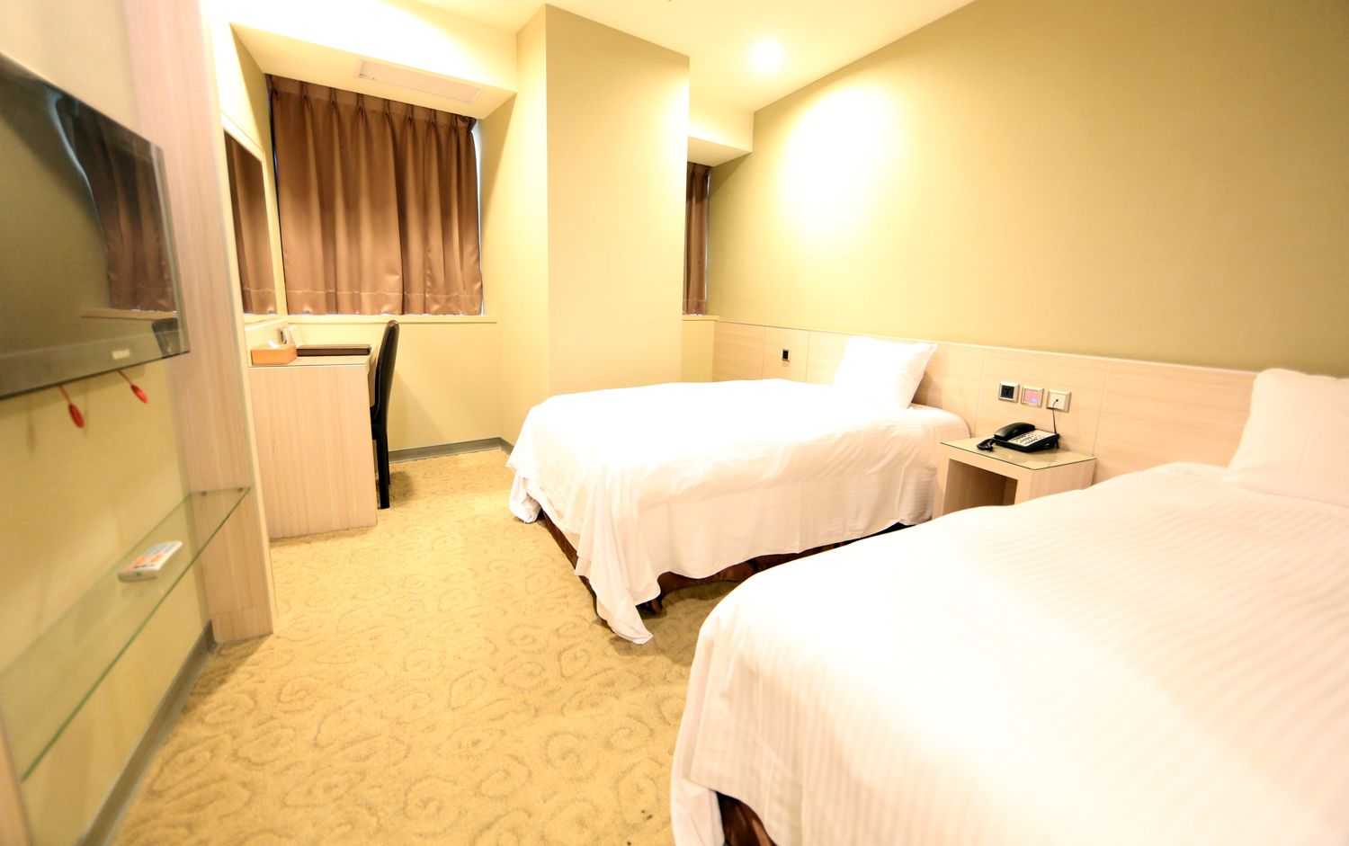 A hotel room with two single beds, a desk with a telephone, a small table, a mirror, and a window with curtains.