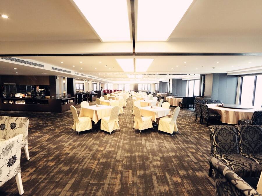 A spacious hotel dining area with round tables and chairs, a bar area, and large windows providing natural light.