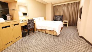 A hotel room with a bed, a desk, a chair, a TV, and a trash can.