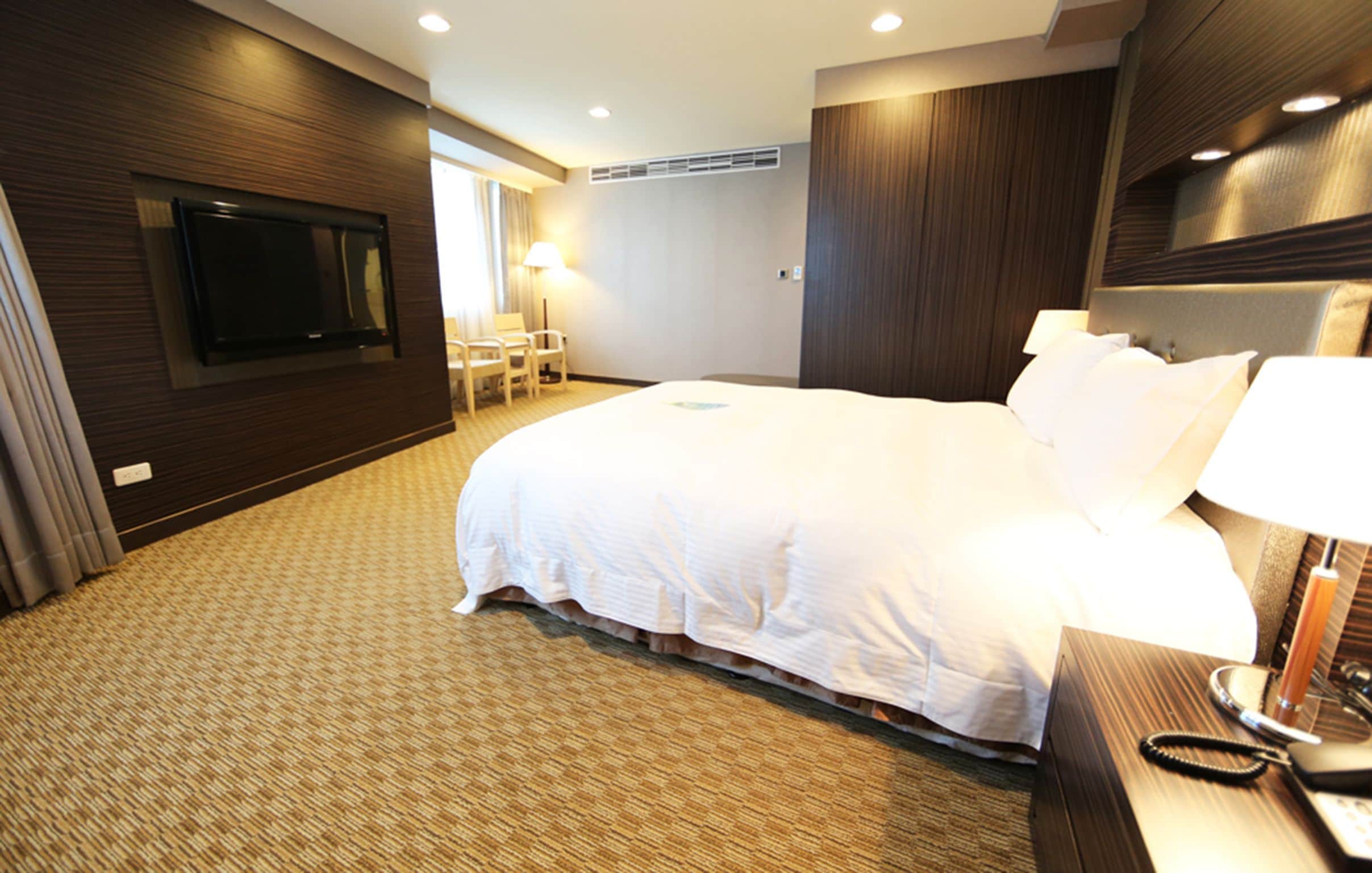 Quarto de hotel com uma cama grande, televisão na parede, mesa de trabalho e telefone, além de uma cadeira.