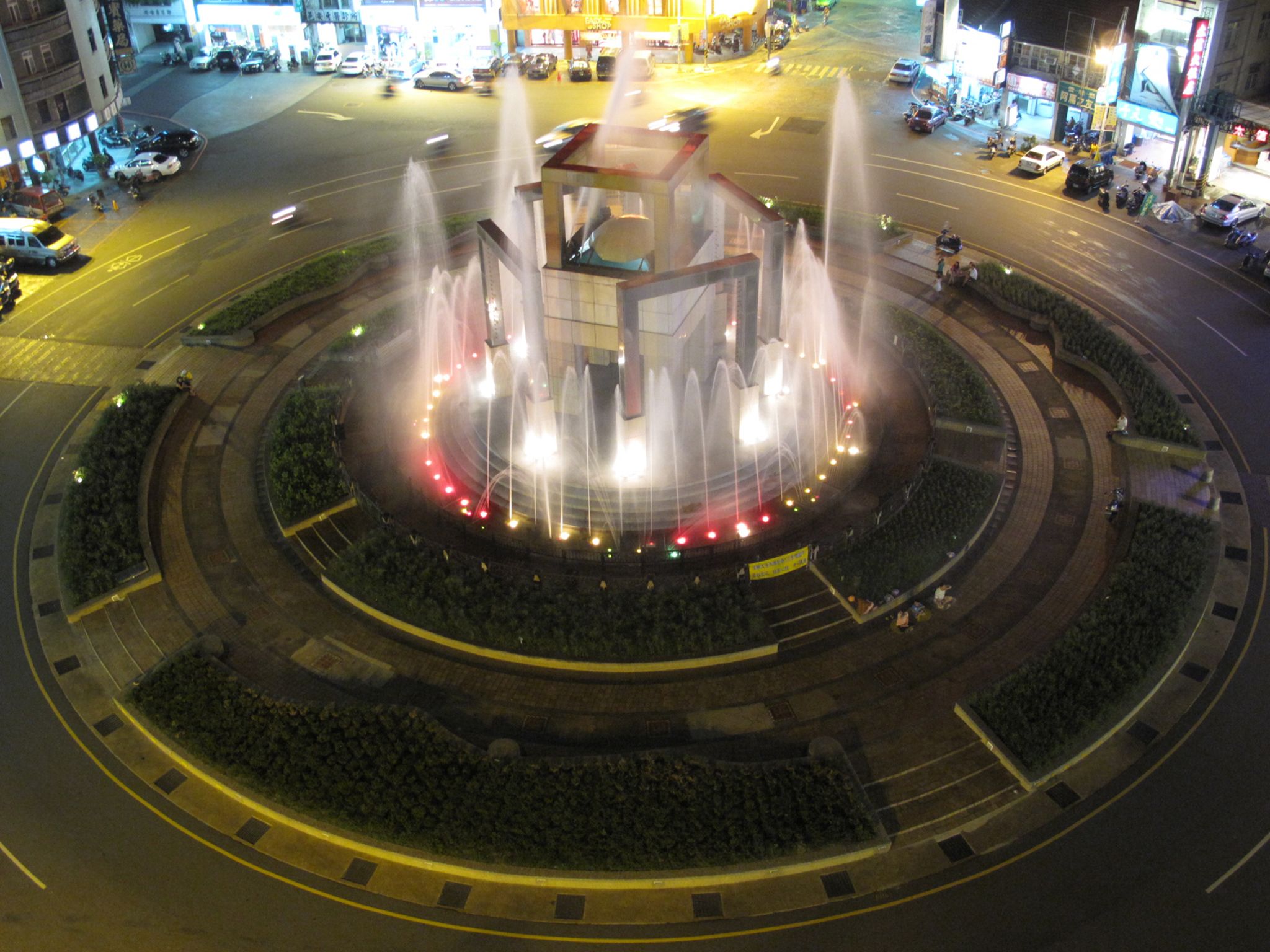 Una fuente circular con múltiples chorros de agua, rodeada de un área ajardinada con bancos y una estructura central.