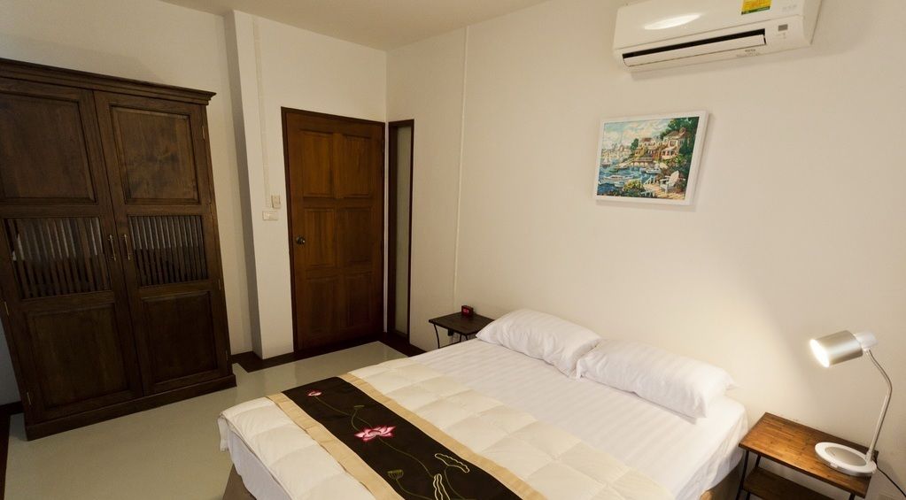 A bedroom with a bed, a wooden wardrobe, a door, a nightstand with a lamp, and a wall-mounted air conditioner.