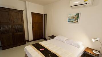 A bedroom with a bed, a wooden wardrobe, a door, a nightstand with a lamp, and a wall-mounted air conditioner.