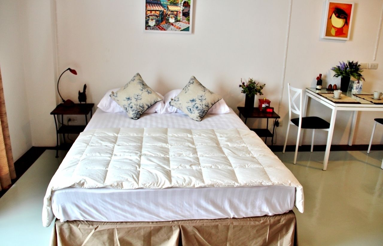 Una cama impecable con un edredón blanco y dos almohadas decorativas, una mesita con un florero y una lámpara, y una silla en un escritorio con una planta.