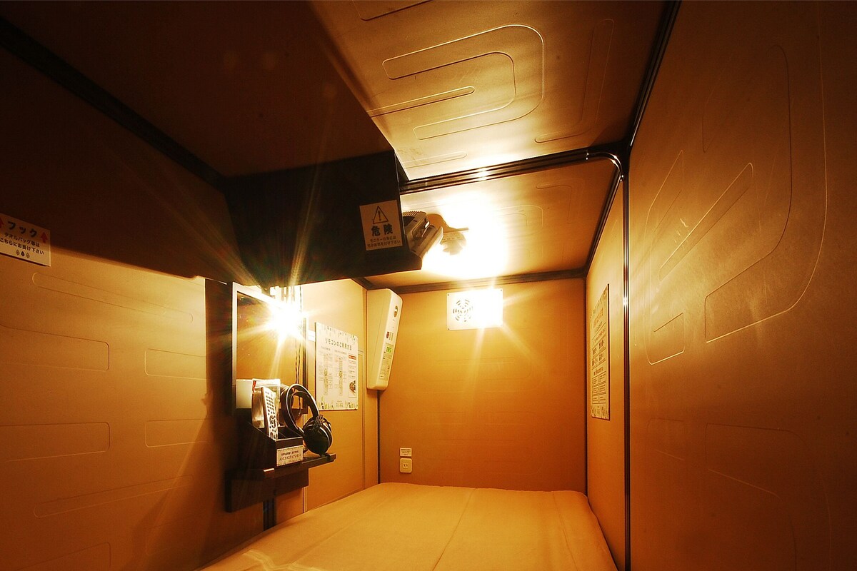 Una habitación estrecha y poco iluminada con una cama, un estante pequeño con auriculares y un espejo.
