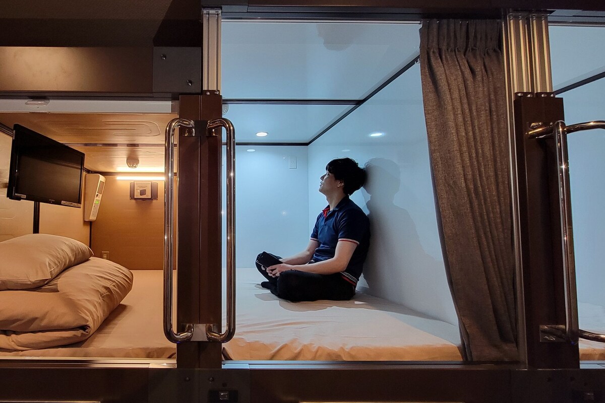 Una habitación compacta y moderna con una cama individual, un televisor montado en la pared y una mesita de noche pequeña.