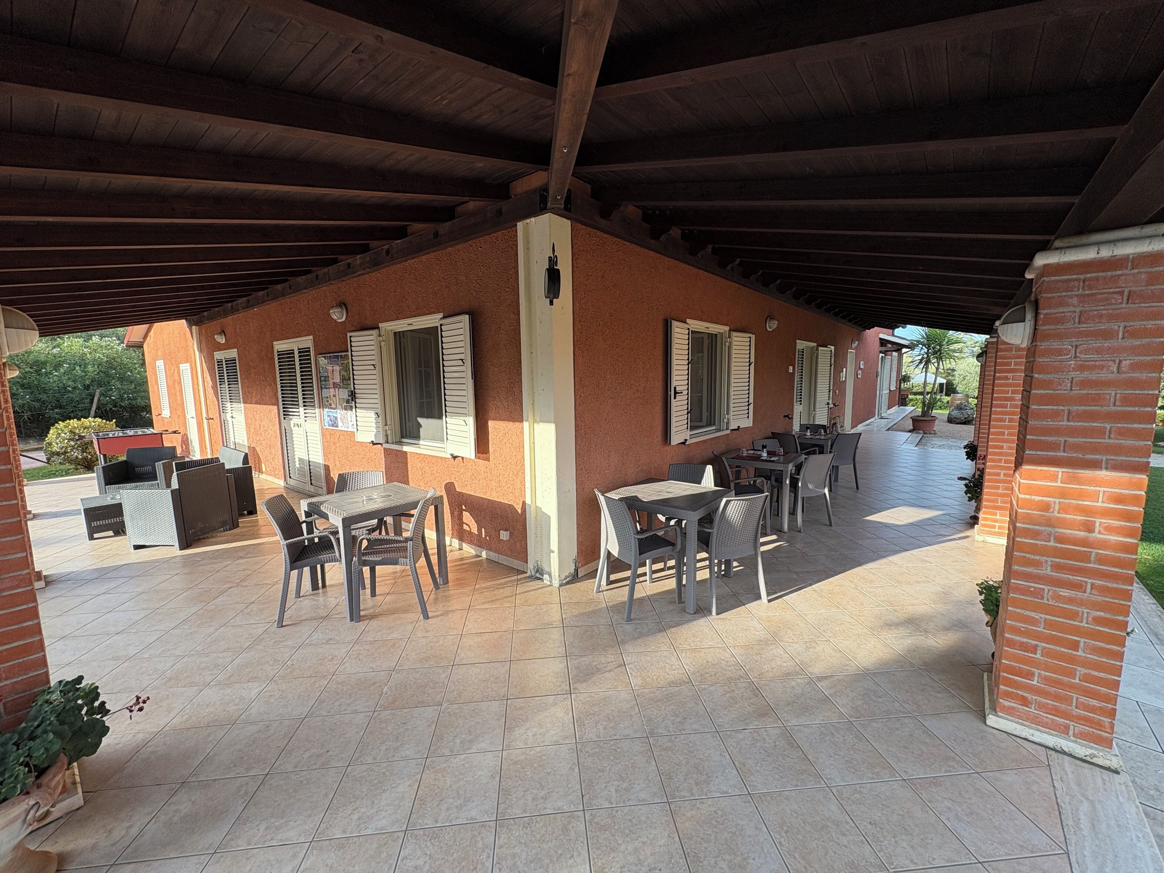 An outdoor patio with brick walls, wooden beams, and tiled flooring. There are several tables and chairs arranged for dining or socializing.