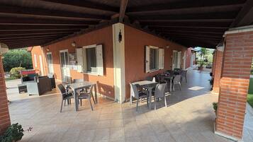 An outdoor patio with brick walls, wooden beams, and tiled flooring. There are several tables and chairs arranged for dining or socializing.