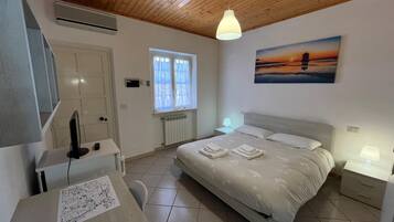 Un dormitorio con cama, mesitas de noche, un escritorio con computadora y una ventana, además de un cuadro en la pared.