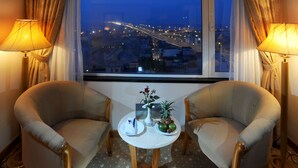 A hotel room with two armchairs, a small table with a vase of flowers, and a view of a cityscape at dusk. - A25 Signature Hotel (Hanoi)