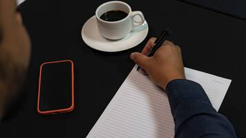 Una persona escribiendo en un cuaderno rayado con una taza de café sobre una mesa.