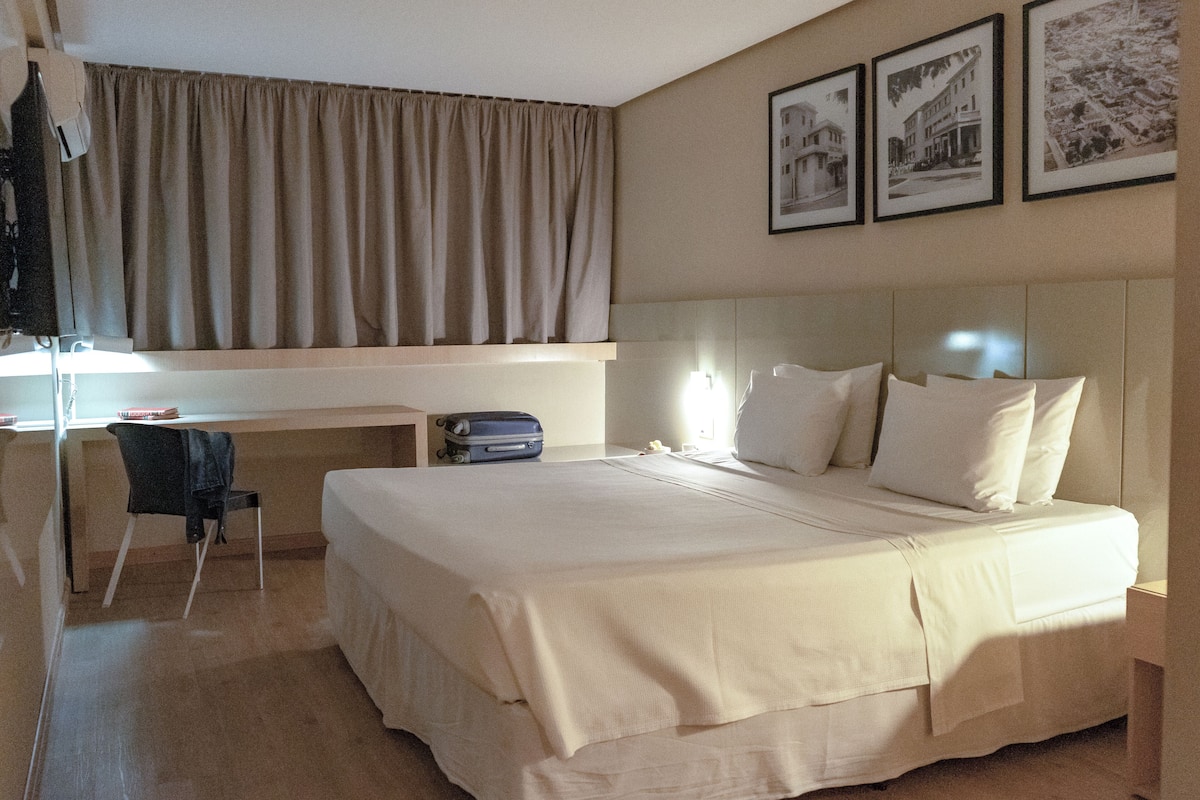 A hotel room with a bed, a desk, a chair, and a window with curtains. There are three framed pictures on the wall.