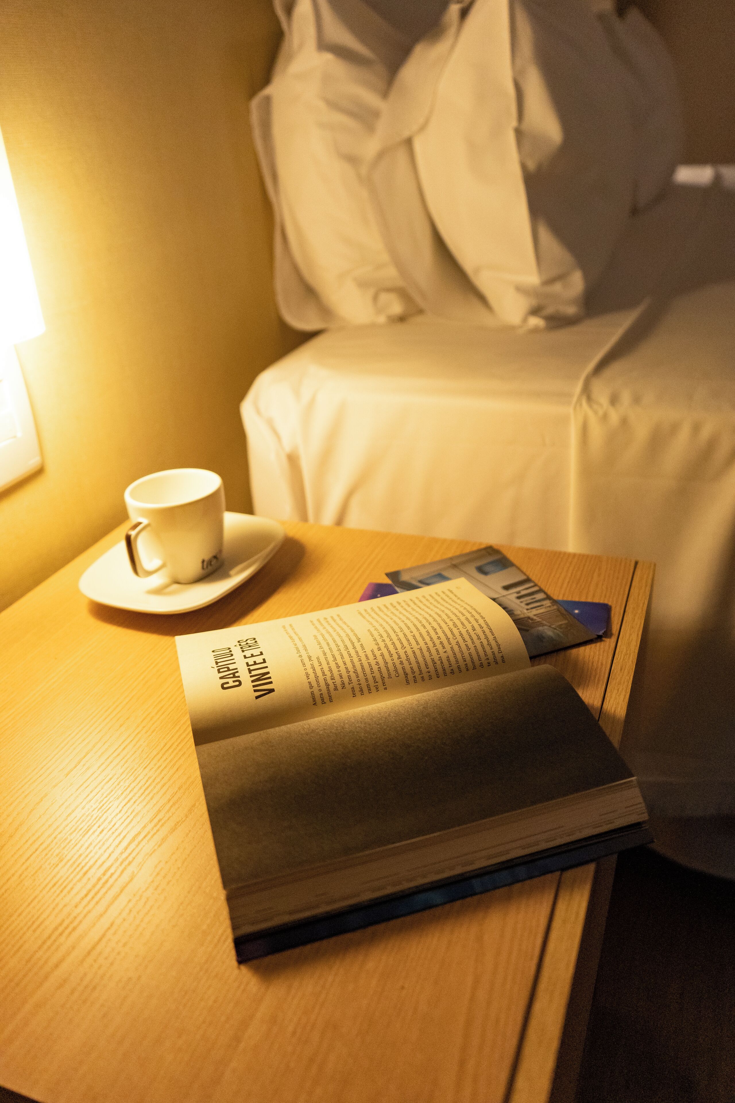 Una cama bien hecha con edredón blanco y almohadas, una mesita de noche con una taza y un plato, y una revista o un libro.