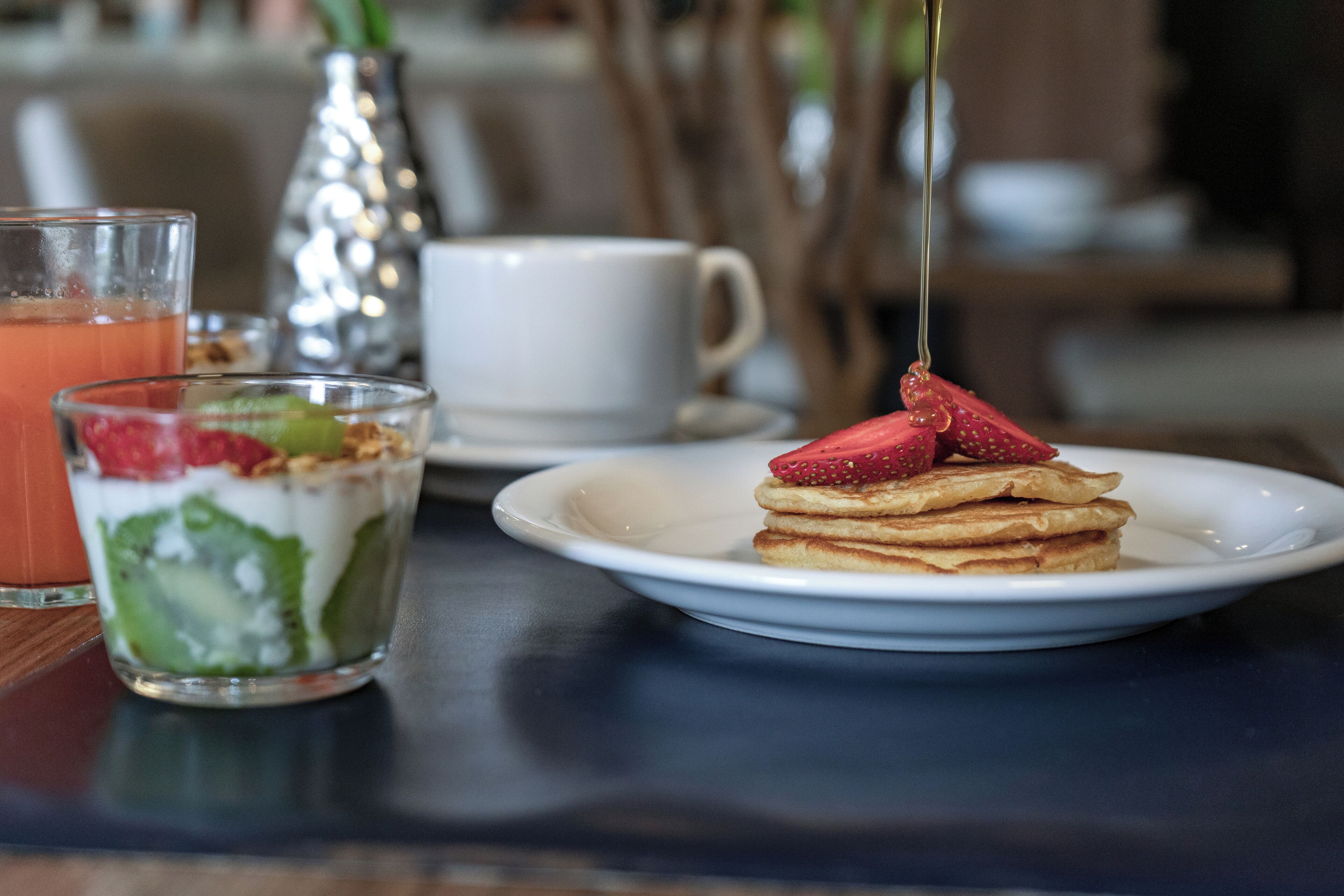 A dessert plate with pancakes and strawberries, a glass of yogurt with fruit, and a white cup on a table.
