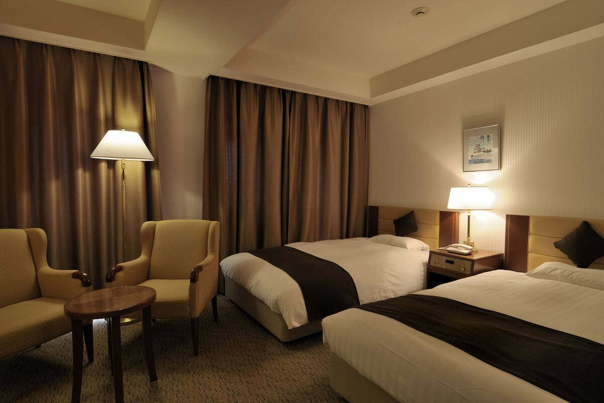 Ein Hotelzimmer mit zwei Betten, einem kleinen Tisch, zwei Sesseln, einer Lampe und einem Nachttisch.