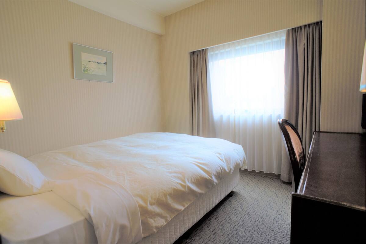 Ein Hotelzimmer mit Bett, Schreibtisch, Lampe und einem Fenster mit Vorhängen.