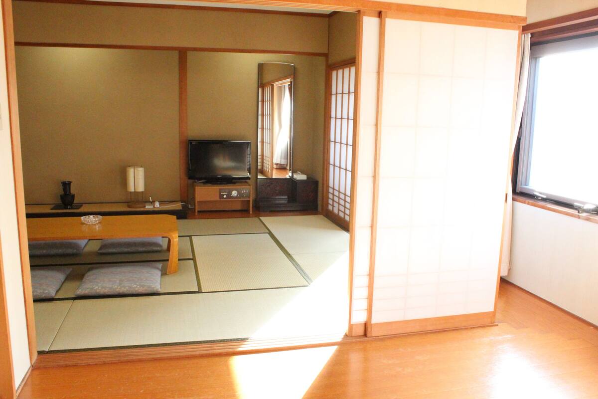 Ein traditioneller japanischer Raum mit Tatami-Böden, einem niedrigen Tisch, einem Fernseher und gleitenden Schiebetüren.