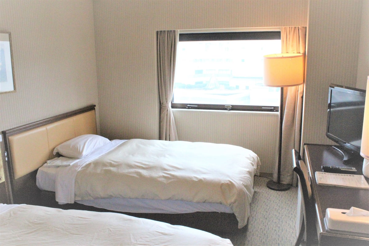Ein Hotelzimmer mit zwei Betten, einem Schreibtisch mit Fernseher und einem Fenster mit Vorhängen.