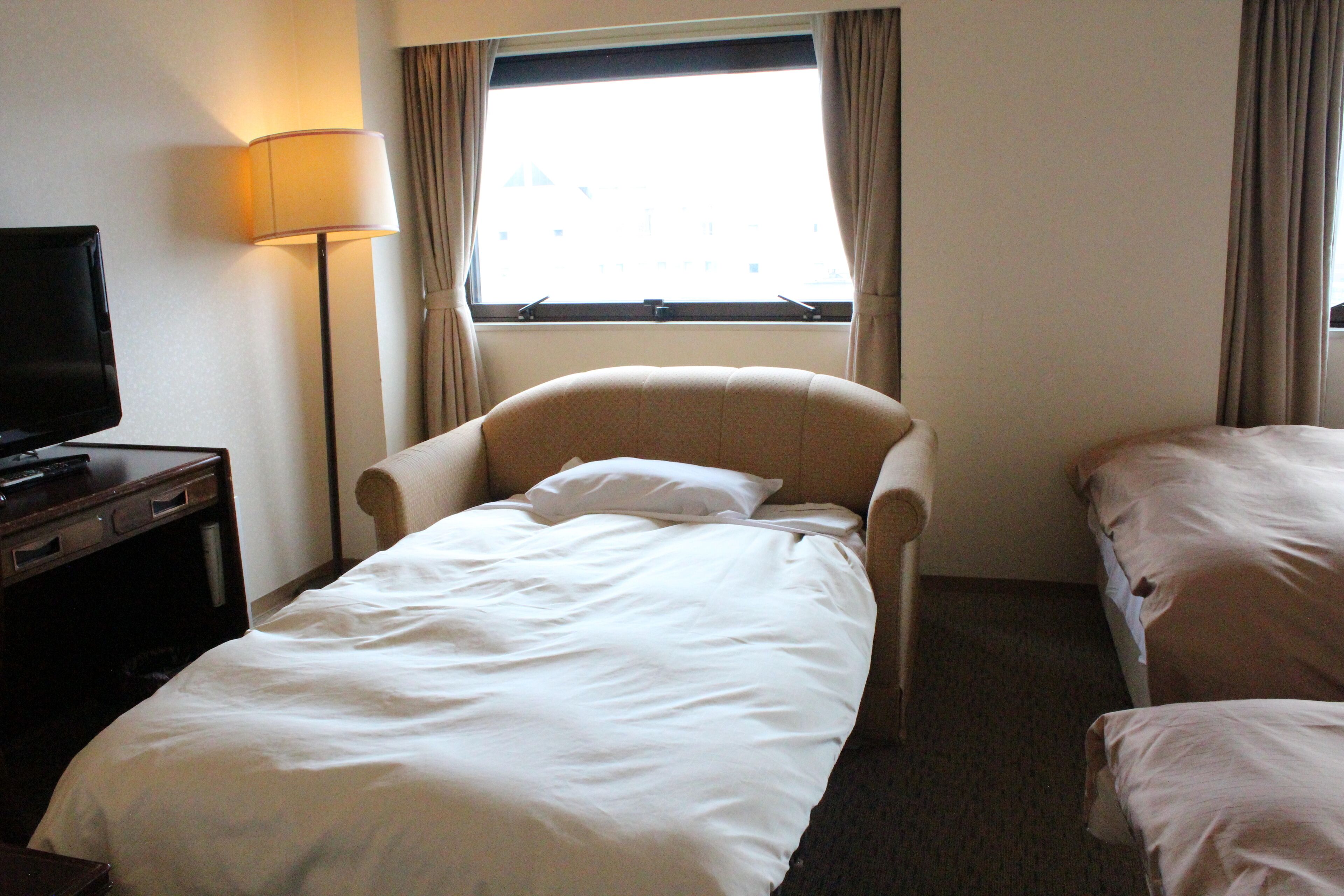 Une chambre d’hôtel avec un lit simple, une chaise, un bureau avec une télévision et une fenêtre avec des rideaux.
