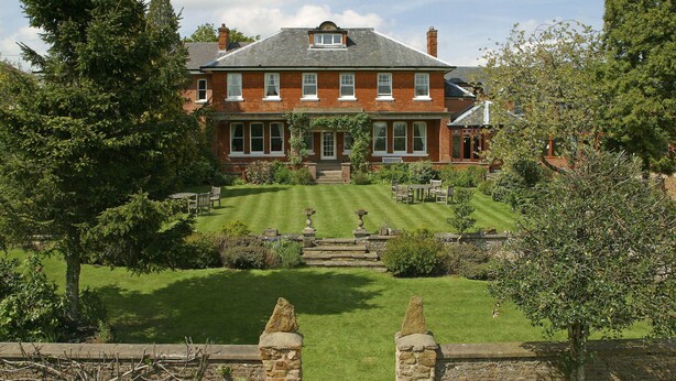 A large brick house with a well-maintained lawn, stone pillars, and a garden with various plants and trees.