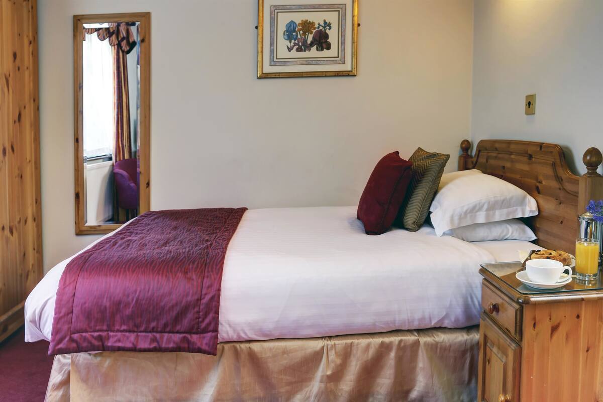 A neatly made bed with a red blanket, a wooden headboard, and a bedside table with a cup of coffee and orange juice.
