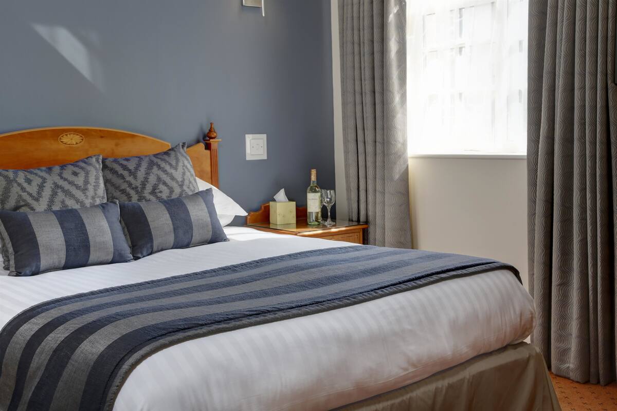A neatly made bed with a striped blanket, a wooden headboard, and patterned pillows. There is a nightstand with a bottle, glass, and tissue box. A window with curtains is visible.