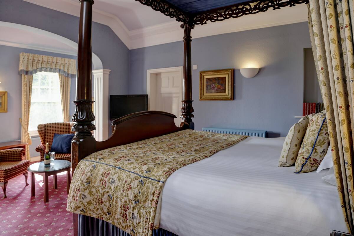 A four-poster bed with a floral bedspread, a television on the wall, and a chair with a bottle of champagne.