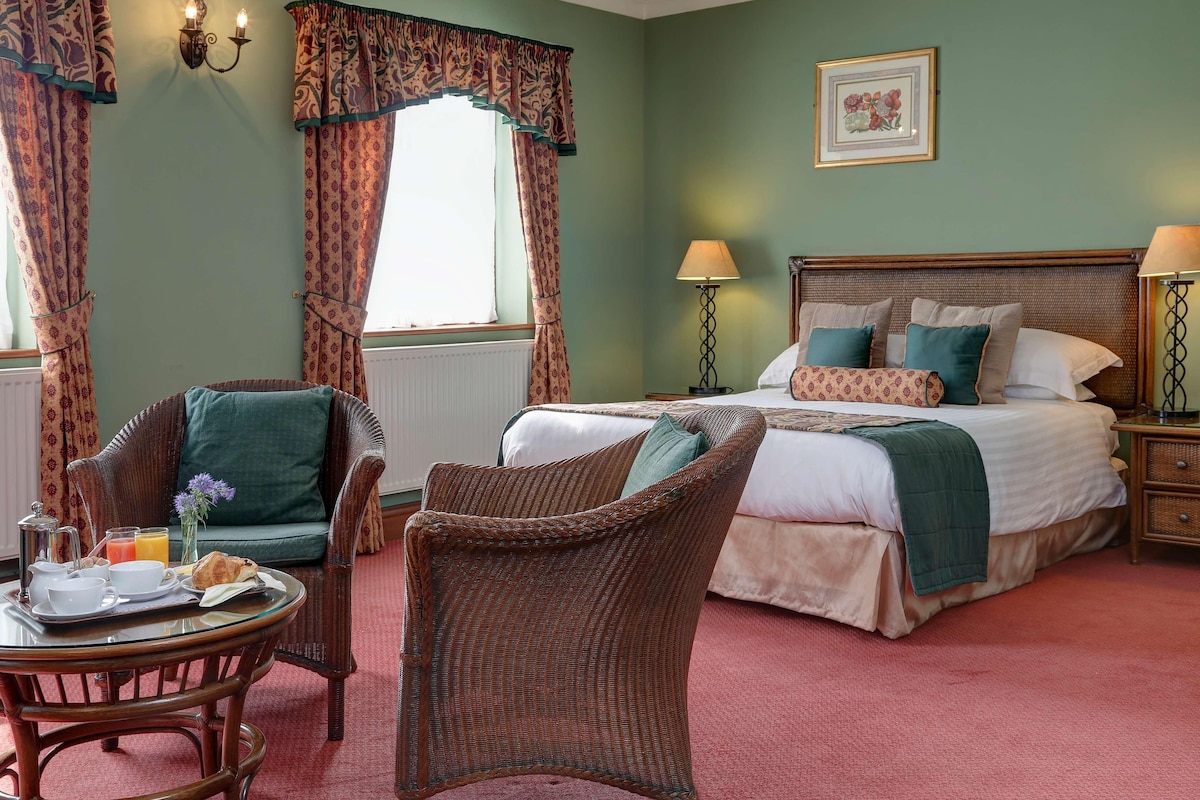 A bedroom with a large bed, two bedside tables with lamps, a wicker chair, a small table with breakfast items, and curtains.