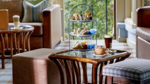 A three-tiered dessert stand with sandwiches and pastries, a coffee cup, and a glass of beer on a table in a cozy room with a sofa and chairs.