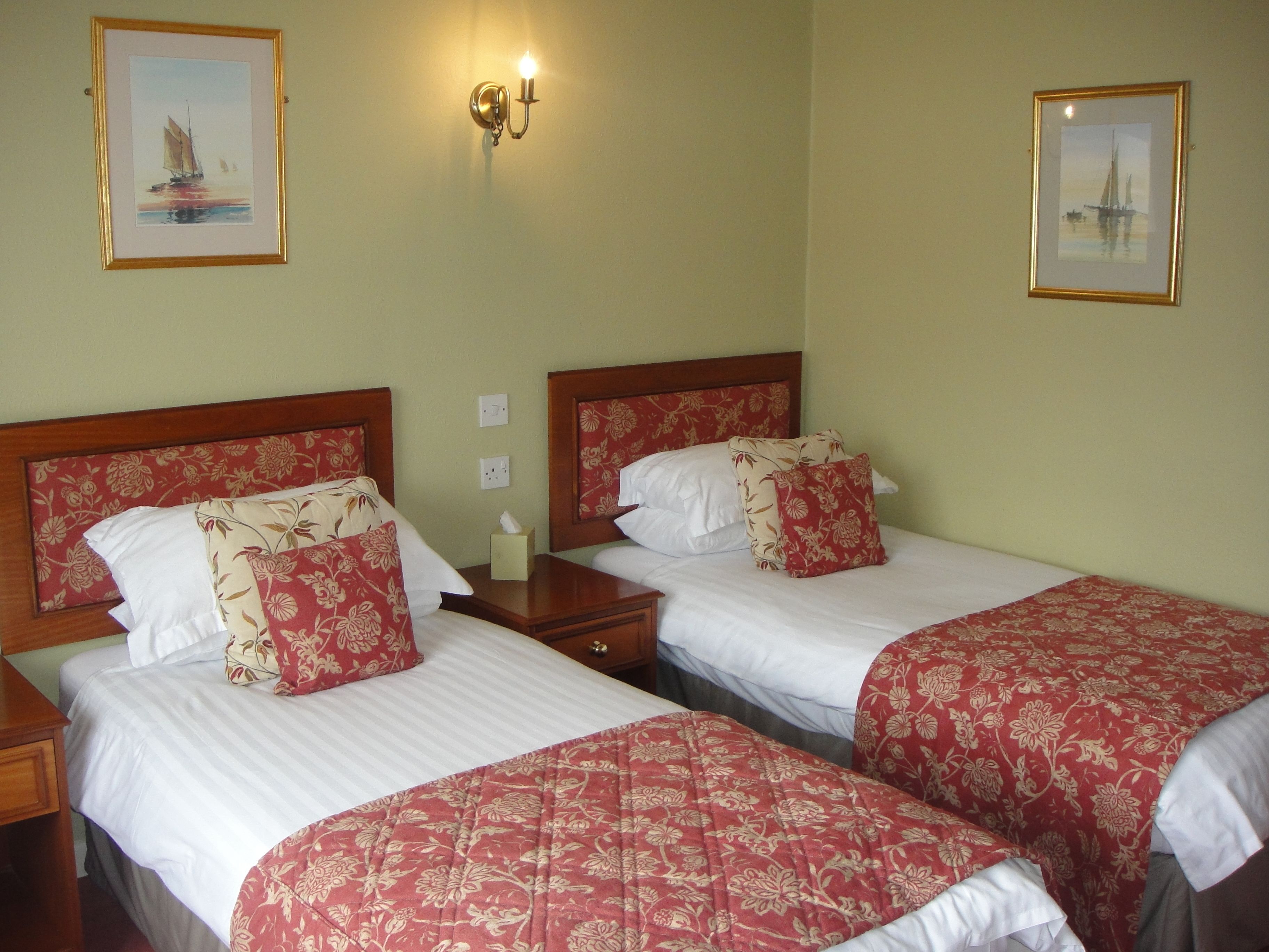 A hotel room with two beds, each with floral-patterned bedding and headboards. There is a wall-mounted light fixture above the beds and two framed pictures on the walls.