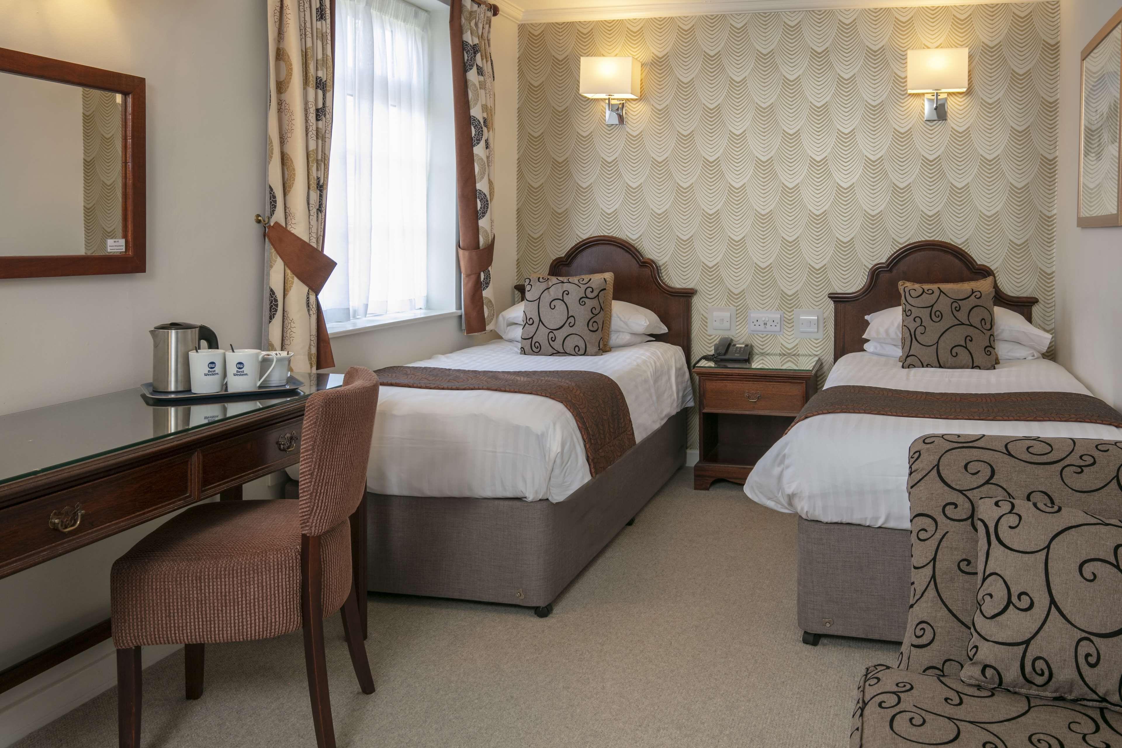 A hotel room with two beds, a desk, a chair, and a lamp.