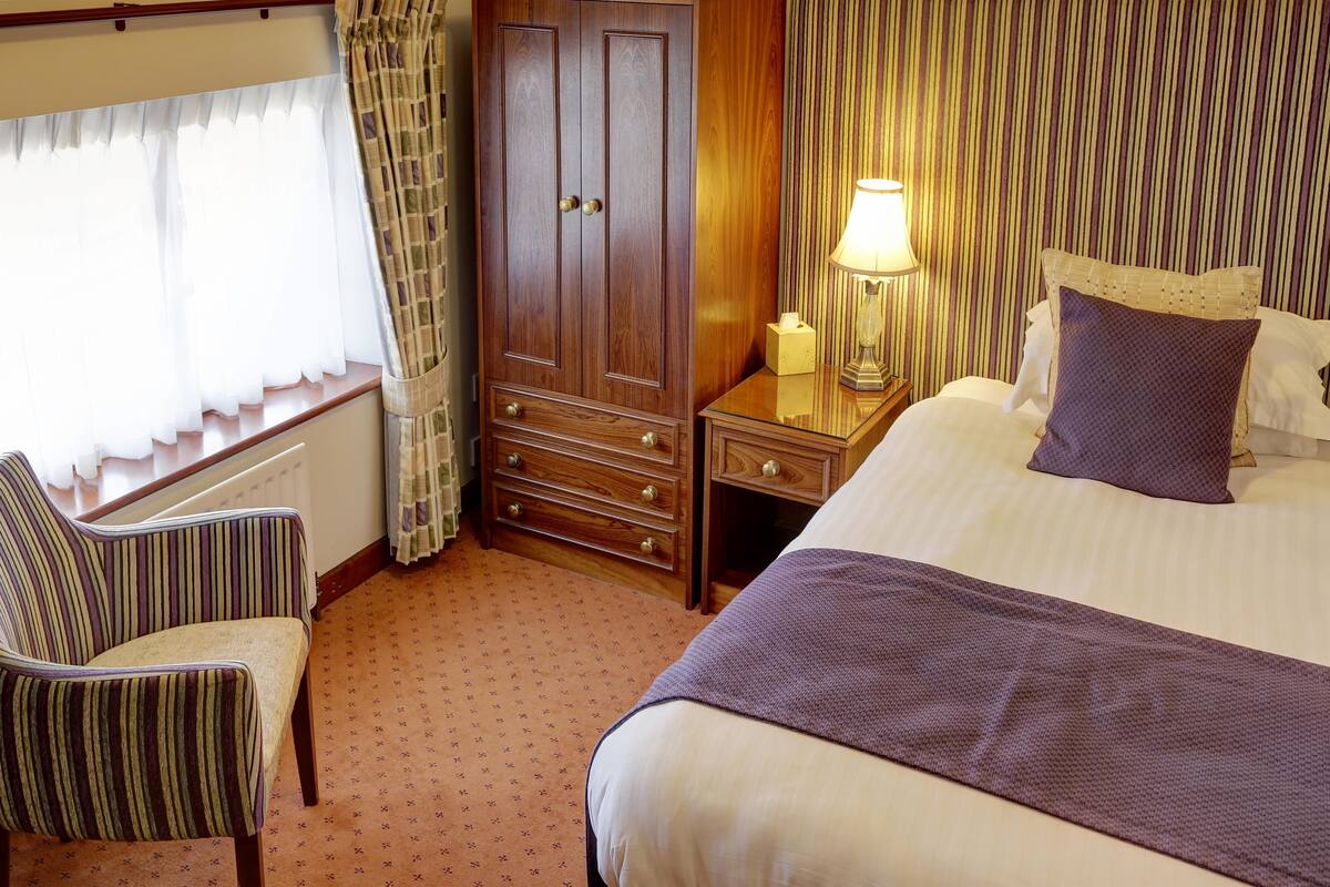 A hotel room with a bed, a chair, a dresser, and a nightstand with a lamp.