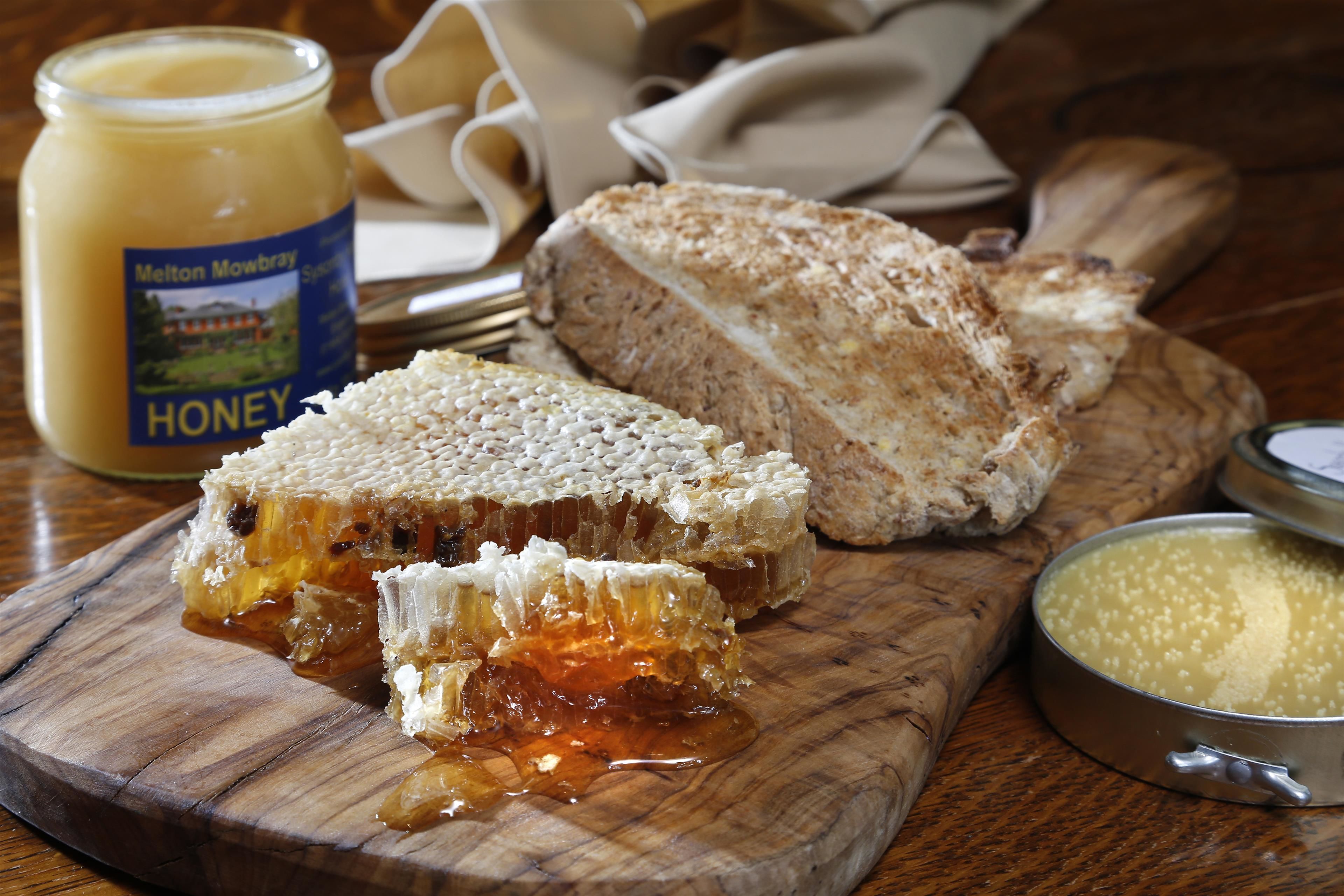 A wooden cutting board with a piece of honeycomb honey, a jar of honey, and a slice of bread.