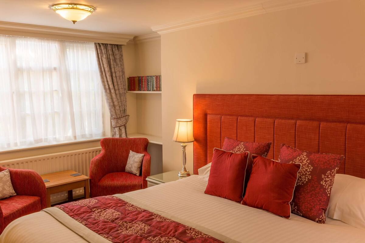 A hotel room with a large bed, two red armchairs, a small table, a lamp, and a bookshelf.