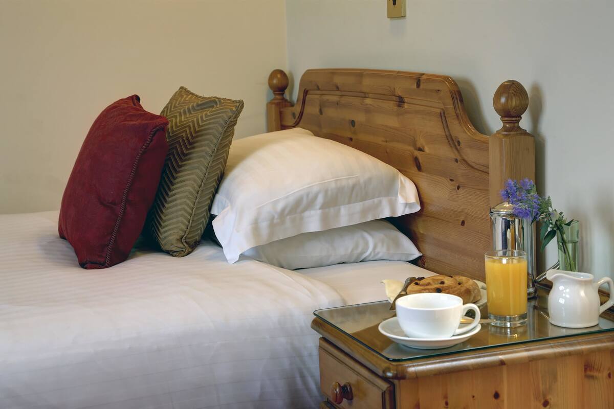 A neatly made bed with white linens and decorative pillows, a bedside table with a glass pitcher of orange juice, a cup, and a plate with cookies.