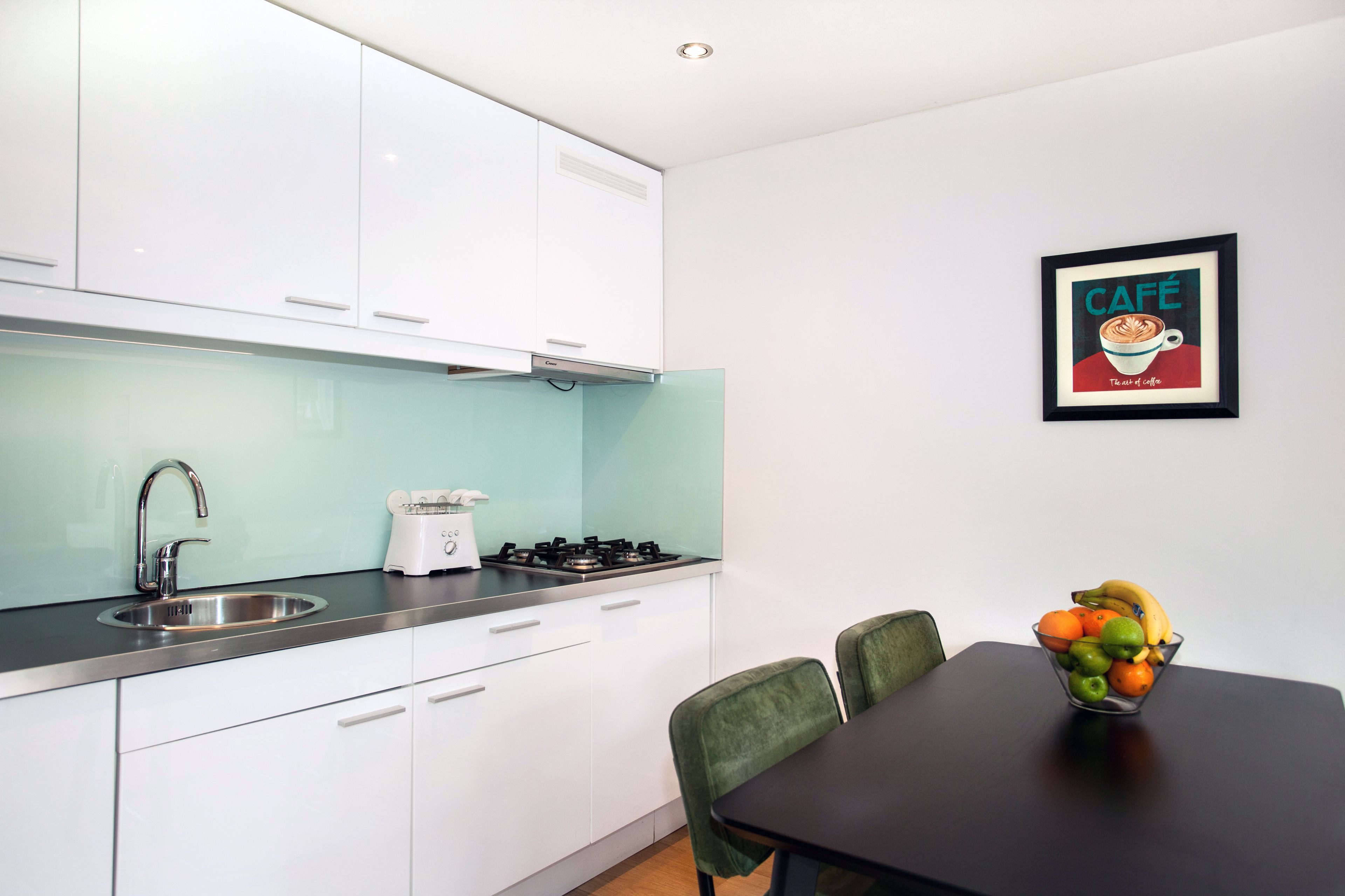 A modern kitchen with white cabinets, a stainless steel sink, and a gas stove. There is a framed picture on the wall and a bowl of fruit on the table.