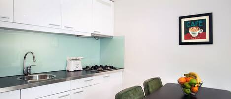 A modern kitchen with white cabinets, a stainless steel sink, and a gas stove. There is a framed picture on the wall and a bowl of fruit on the table.