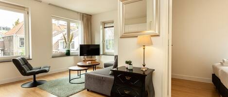 A modern hotel room with a large bed, a desk with a TV, a comfortable chair, and a large mirror.