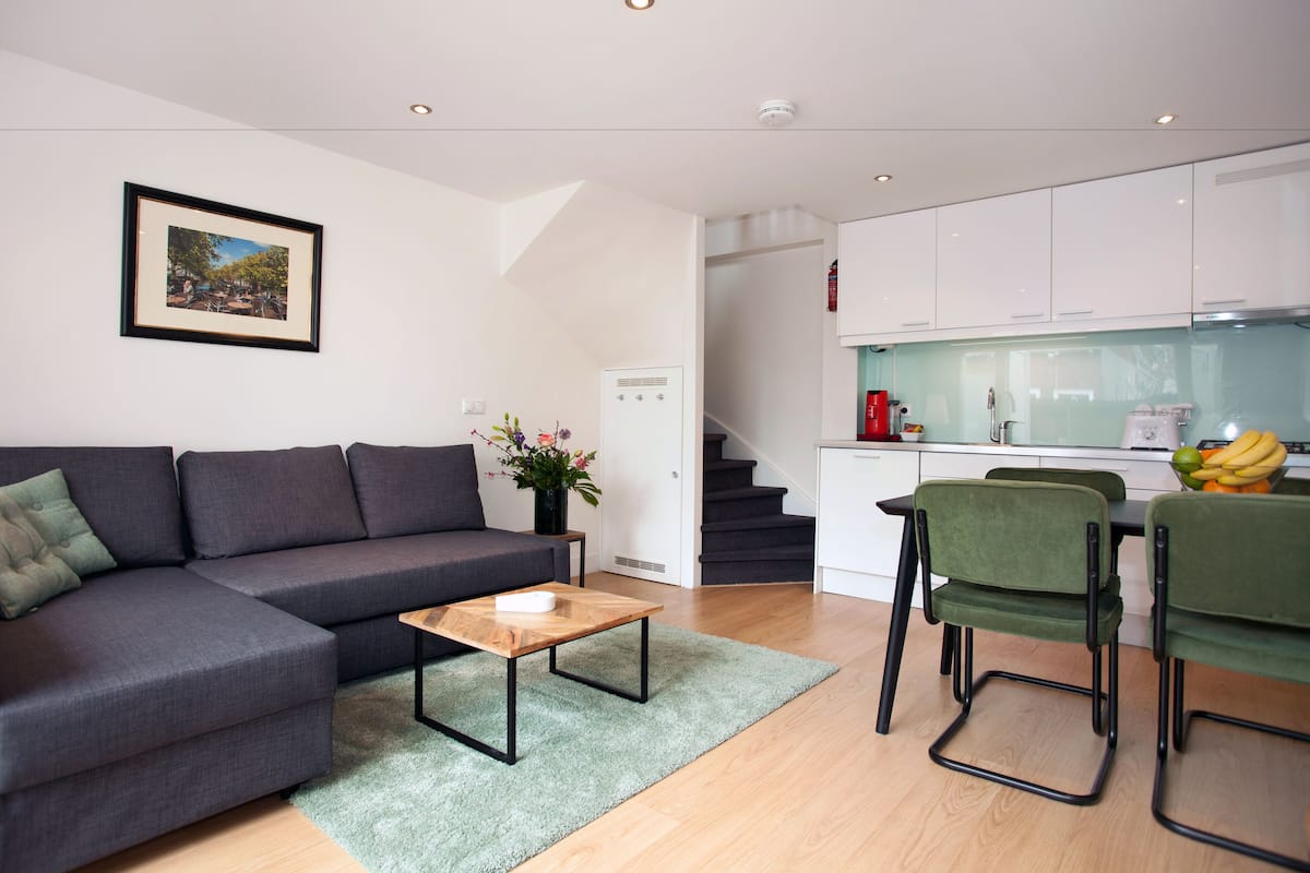 Una sala moderna con un sofá gris, una mesa de centro de madera y una alfombra verde. Hay una zona de cocina con armarios blancos y una mesa de comedor con sillas verdes.
