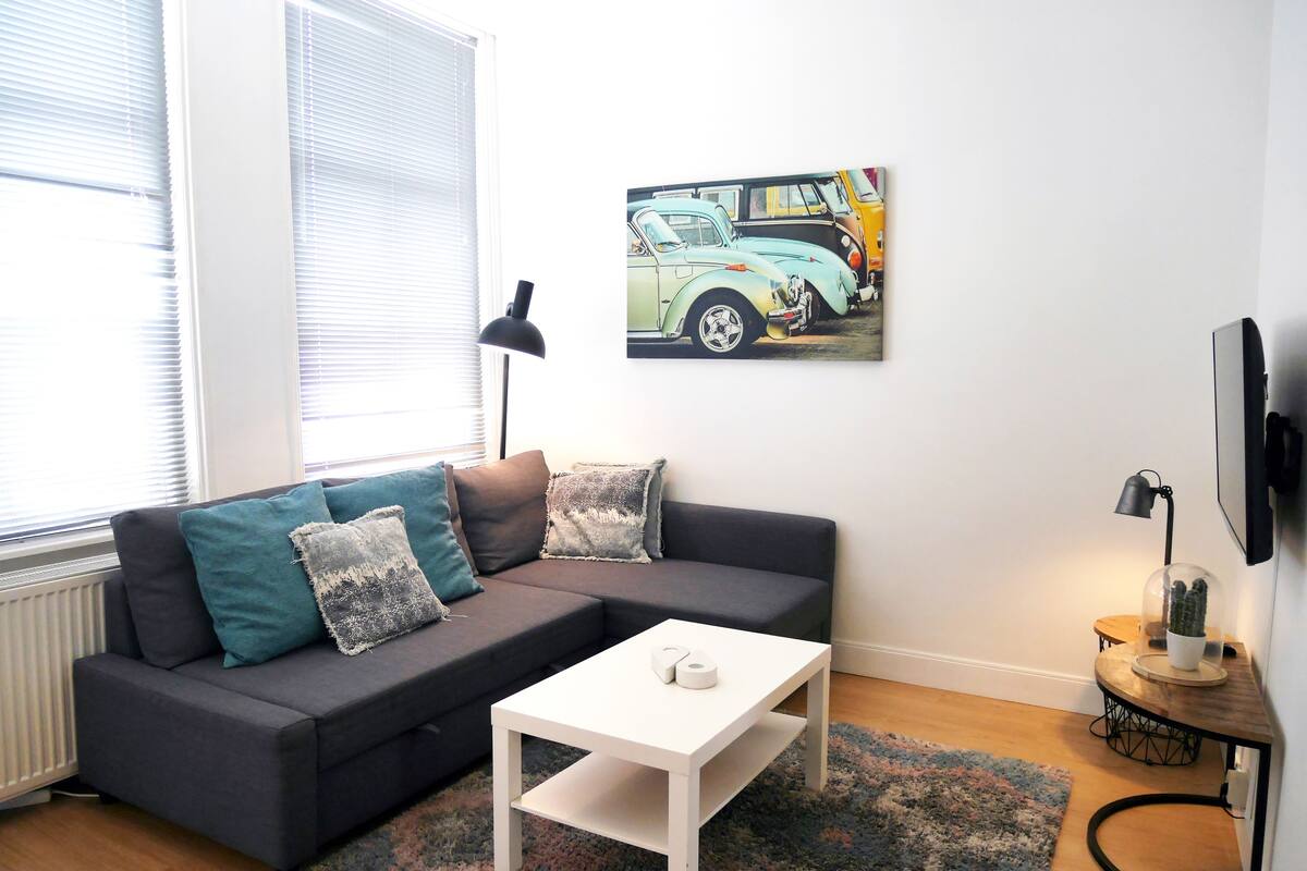 Una sala de estar moderna con un sofá gris oscuro, cojines verde azulado y gris, una mesa de centro blanca y un cuadro con coches antiguos en la pared.