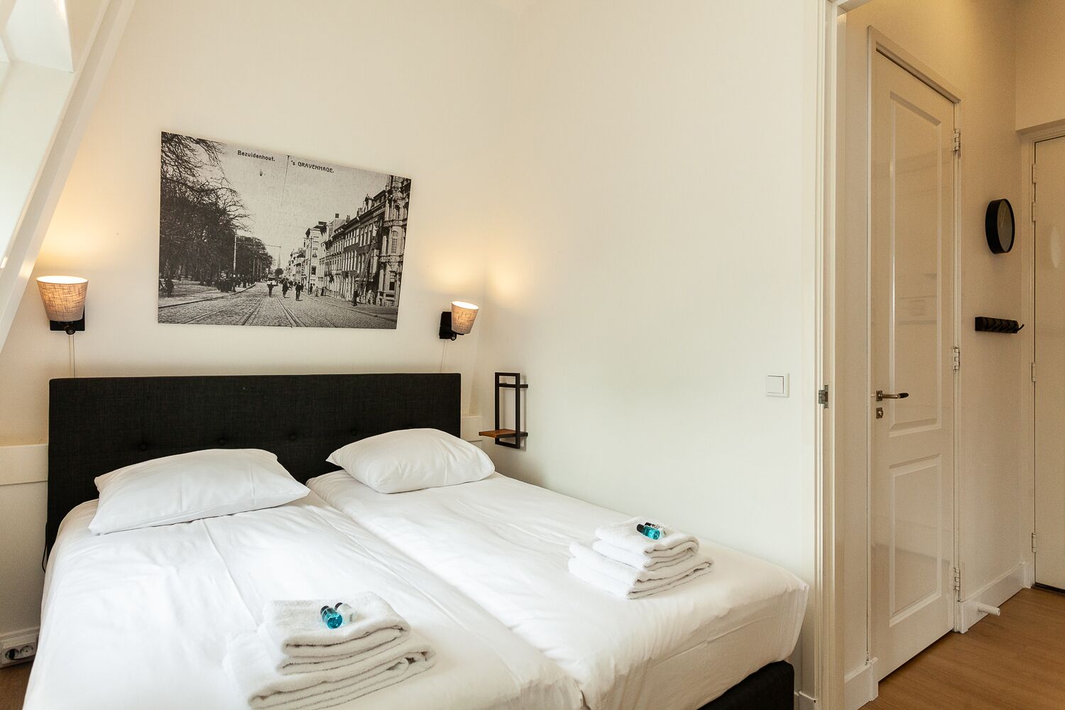 Una cama doble con ropa de cama blanca, dos lámparas fijadas a la pared, una fotografía en blanco y negro enmarcada y una puerta a la derecha.