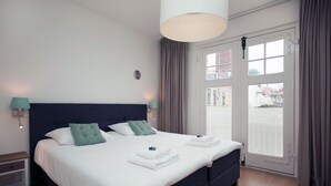 Habitació d'hotel amb un llit gran, dues taules de nit, una finestra amb cortines i vistes a edificis.
