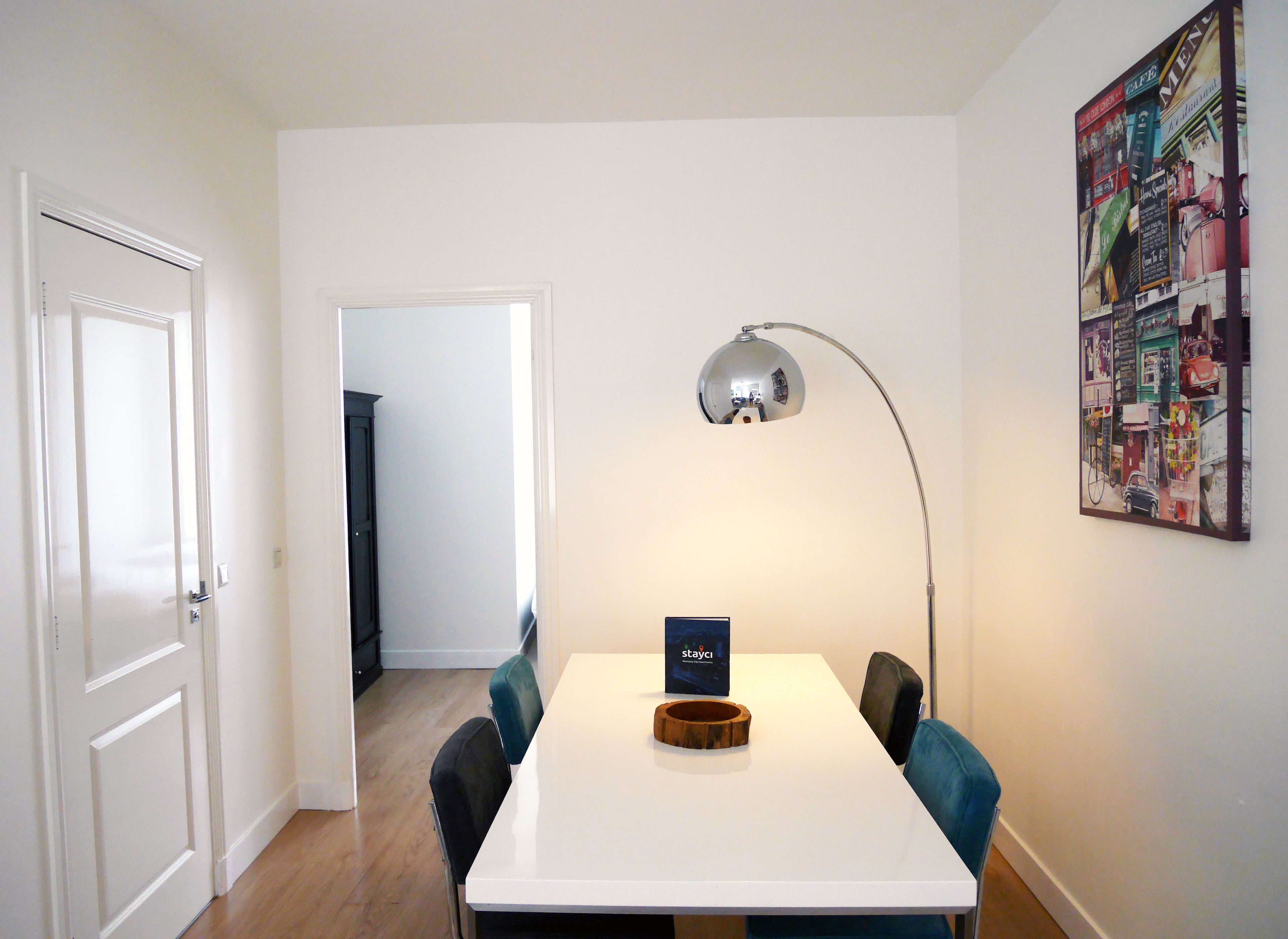 A dining area with a white table, two blue chairs, a wooden bowl, a black sign, and a floor lamp.