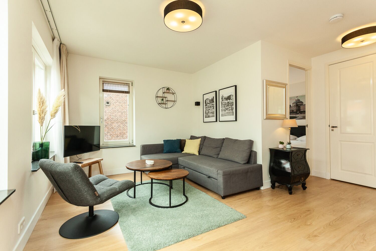 A modern living room with a grey sofa, a round coffee table, a grey armchair, a TV, and a window with a view of a brick building.
